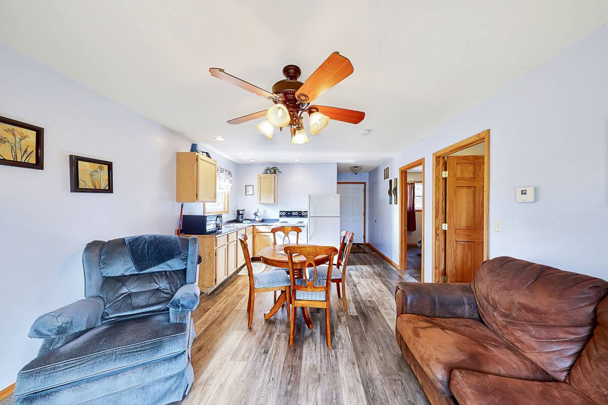 A living room with a sofa, a brown leather couch, a dining table with chairs, a ceiling fan, and a kitchen area in the background.