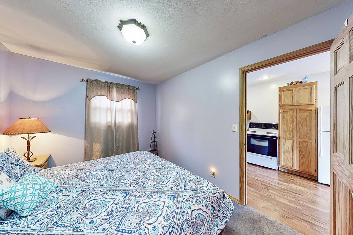 A bedroom with a patterned bedspread, a bedside lamp, and a view into the kitchen through an open door.