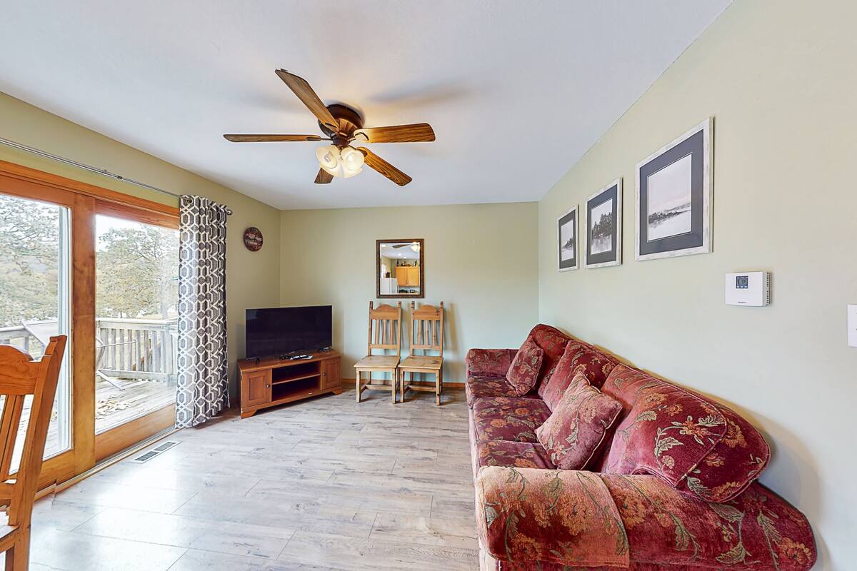 A living room with a wooden floor, a red sofa, a ceiling fan, a TV, and a balcony with patterned curtains.