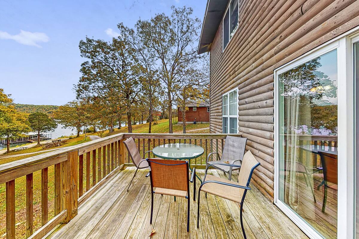 A wooden deck with a glass table and chairs, overlooking a lake and trees.