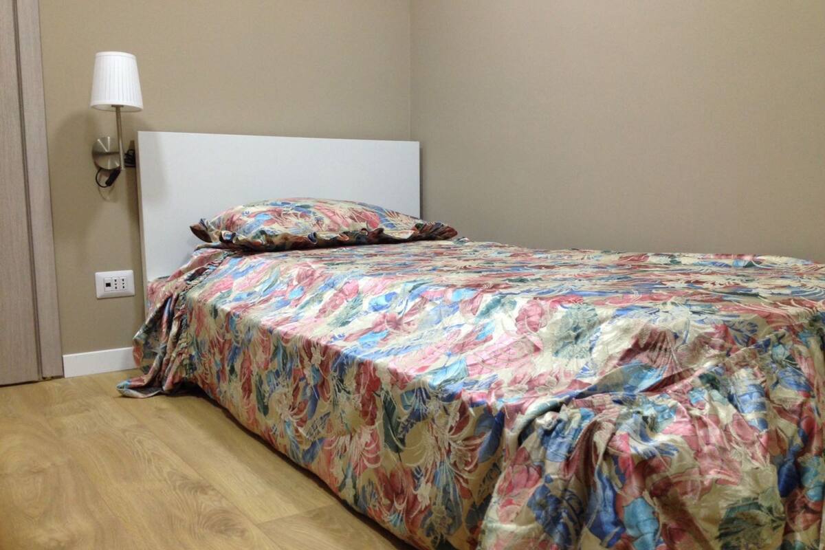 A bed with a floral bedspread, a white headboard, and a wall-mounted lamp.