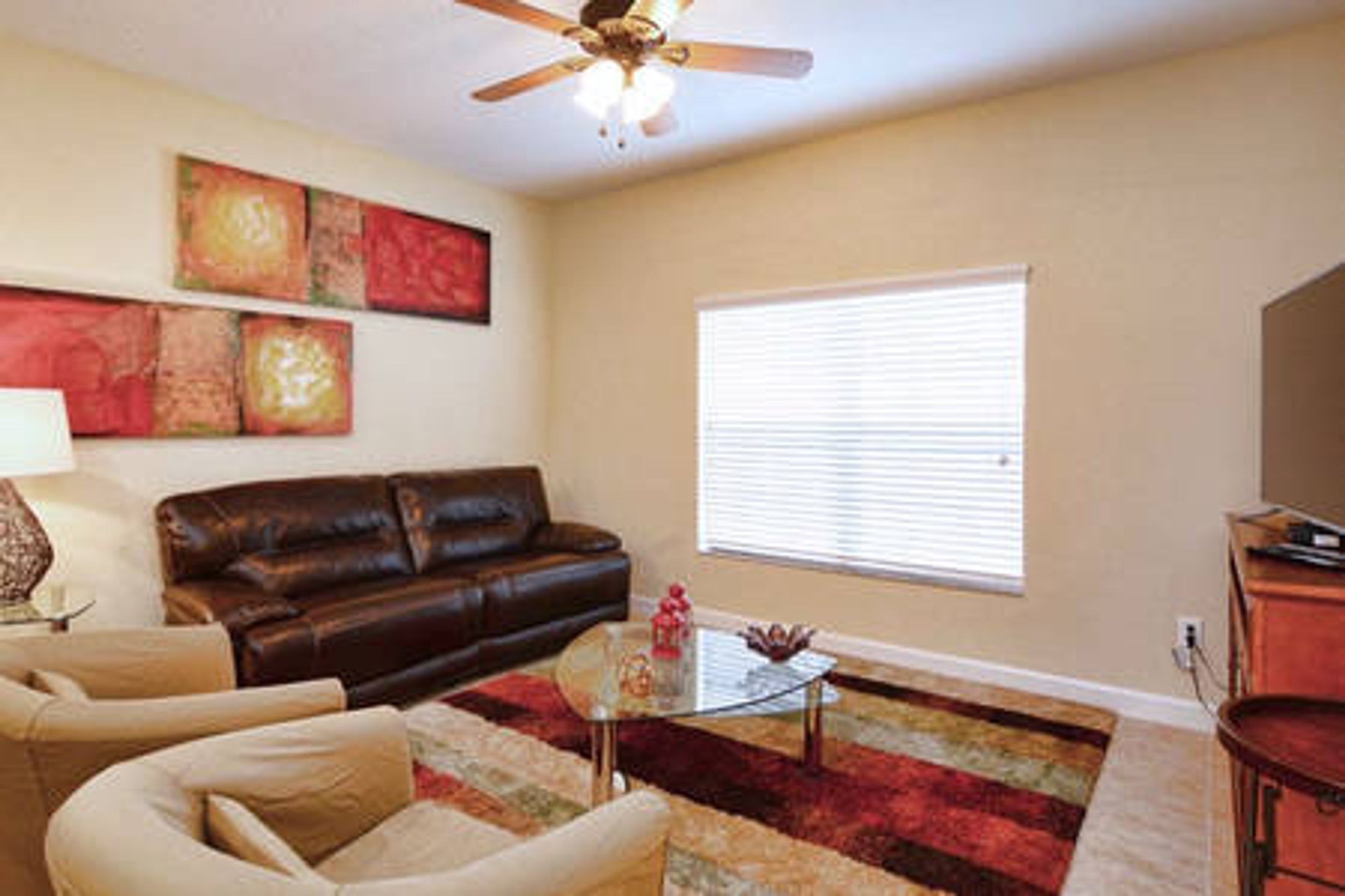 A living room with a brown leather couch, a glass coffee table, a red and beige rug, a ceiling fan, and a window with blinds.