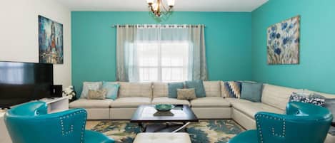 A living room with teal walls, a sofa set, two teal chairs, a coffee table, a TV, and artwork on the walls.