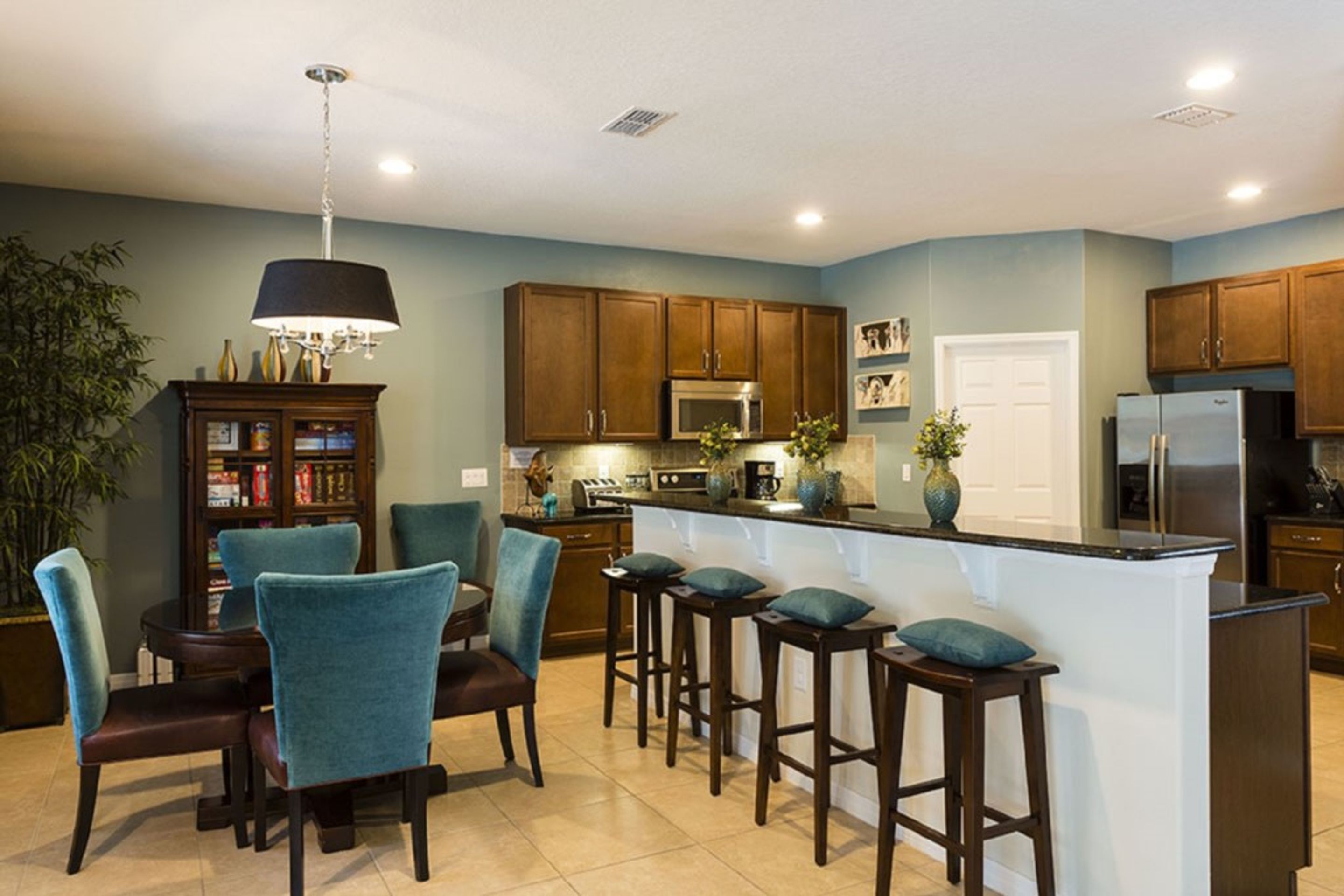 A modern kitchen with dark wooden cabinets, a central island with bar stools, and a dining area with teal chairs.