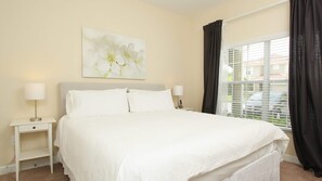 A neatly arranged bedroom with a large bed, two bedside tables with lamps, a window with curtains, and a floral painting on the wall.