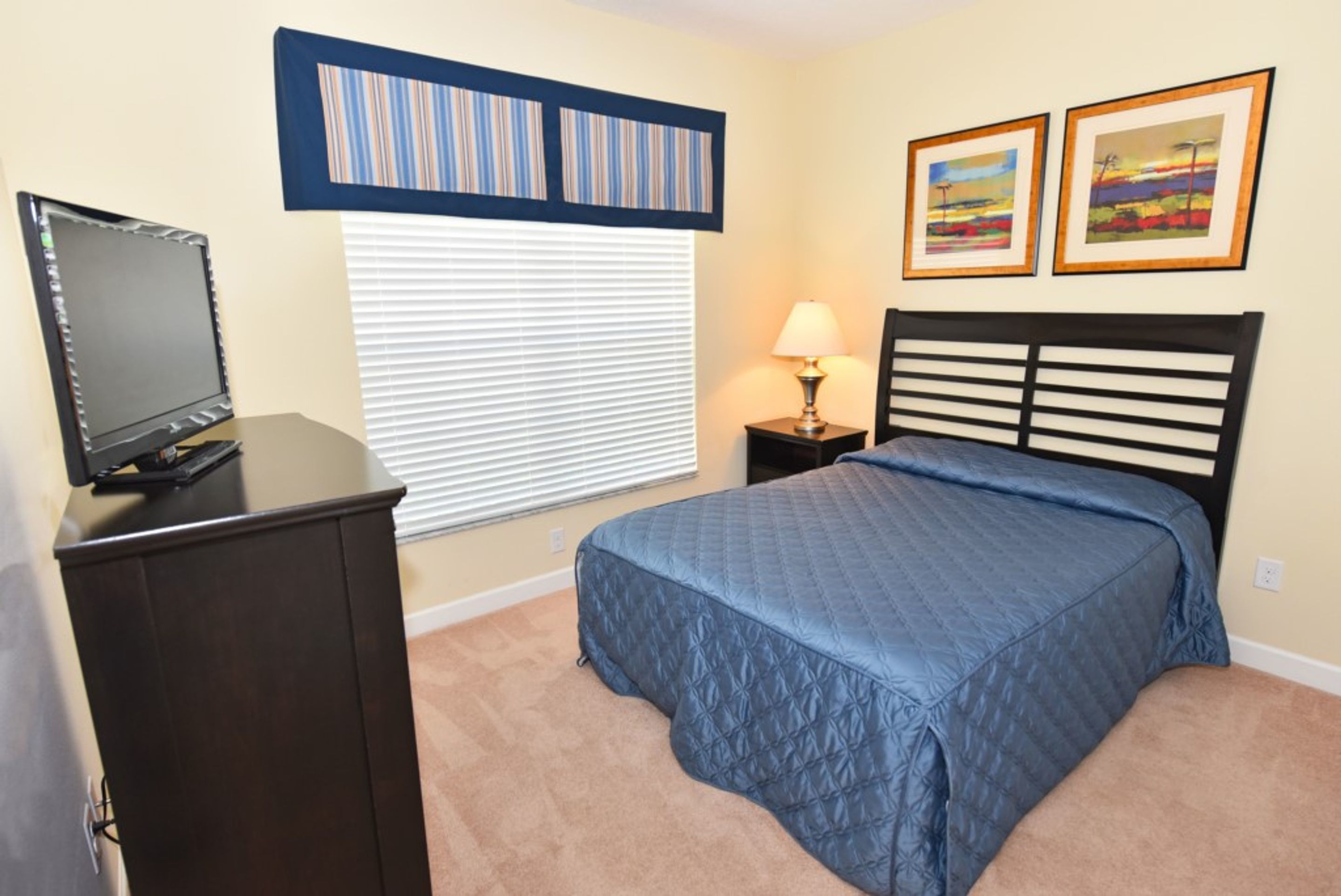 A bedroom with a bed, a desk with a computer, a lamp, and two framed pictures on the wall.