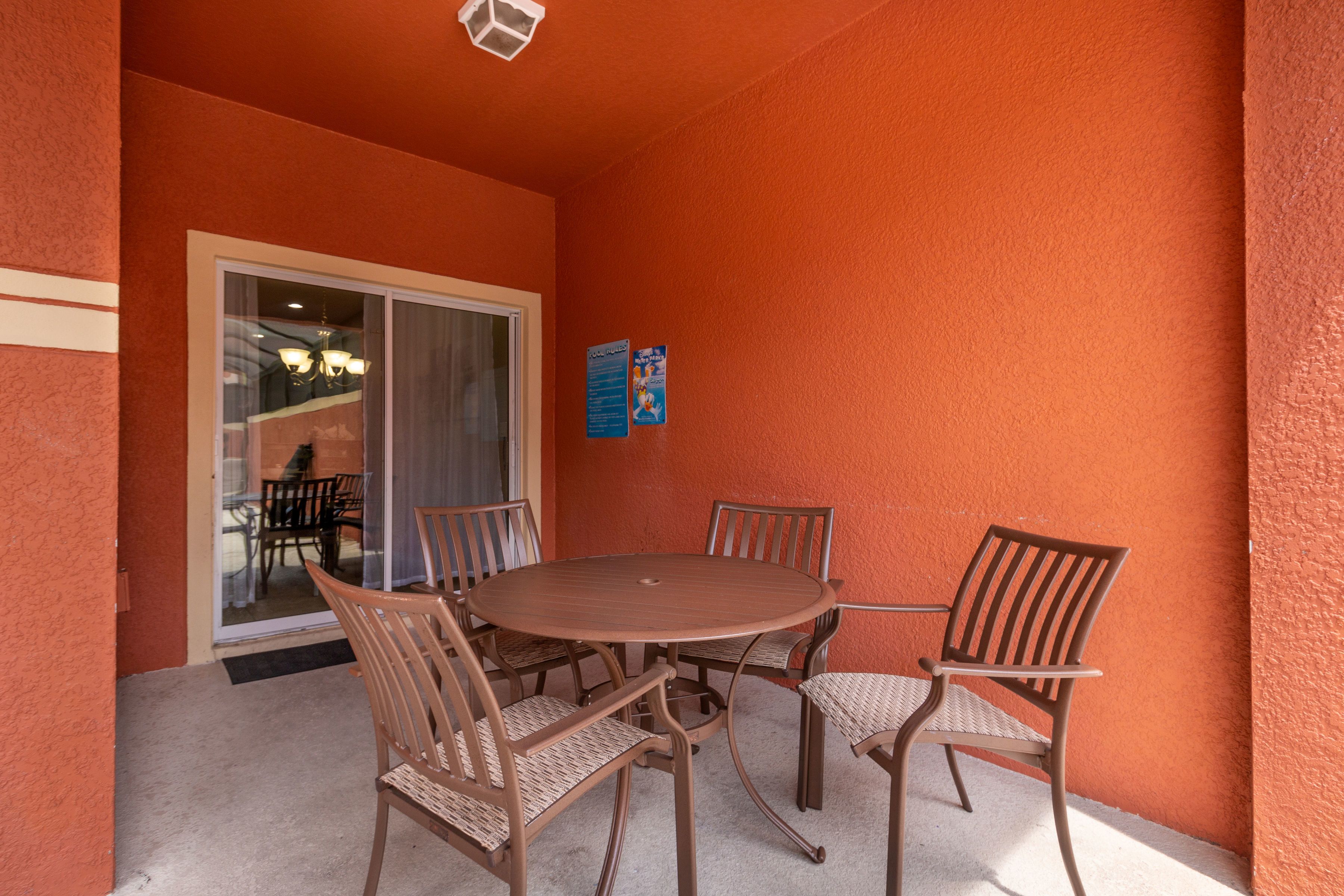 Une table ronde entourée de six chaises, située dans un espace extérieur avec un mur orange et une porte vitrée donnant sur un espace intérieur.