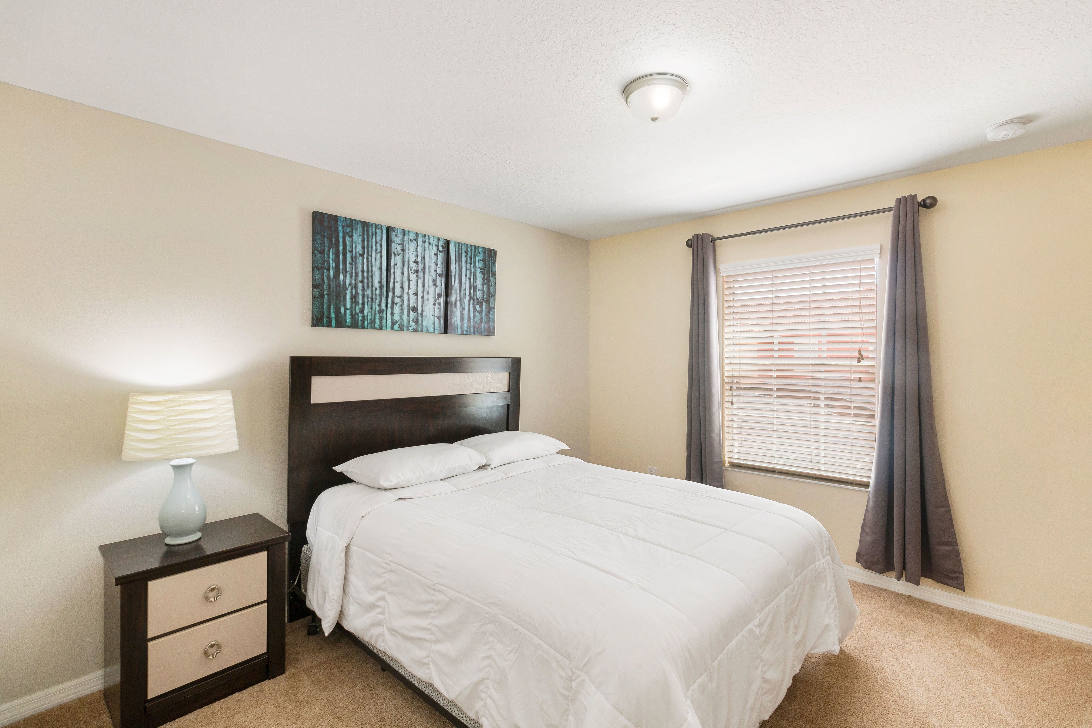 A bedroom with a bed, a nightstand with drawers, a lamp, a ceiling light, a window with curtains, and a wall art piece.