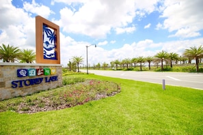 A sign for Story Lake with a palm tree and beach scene, surrounded by green grass and palm trees. - Storey Lake 3131 (Kissimmee)