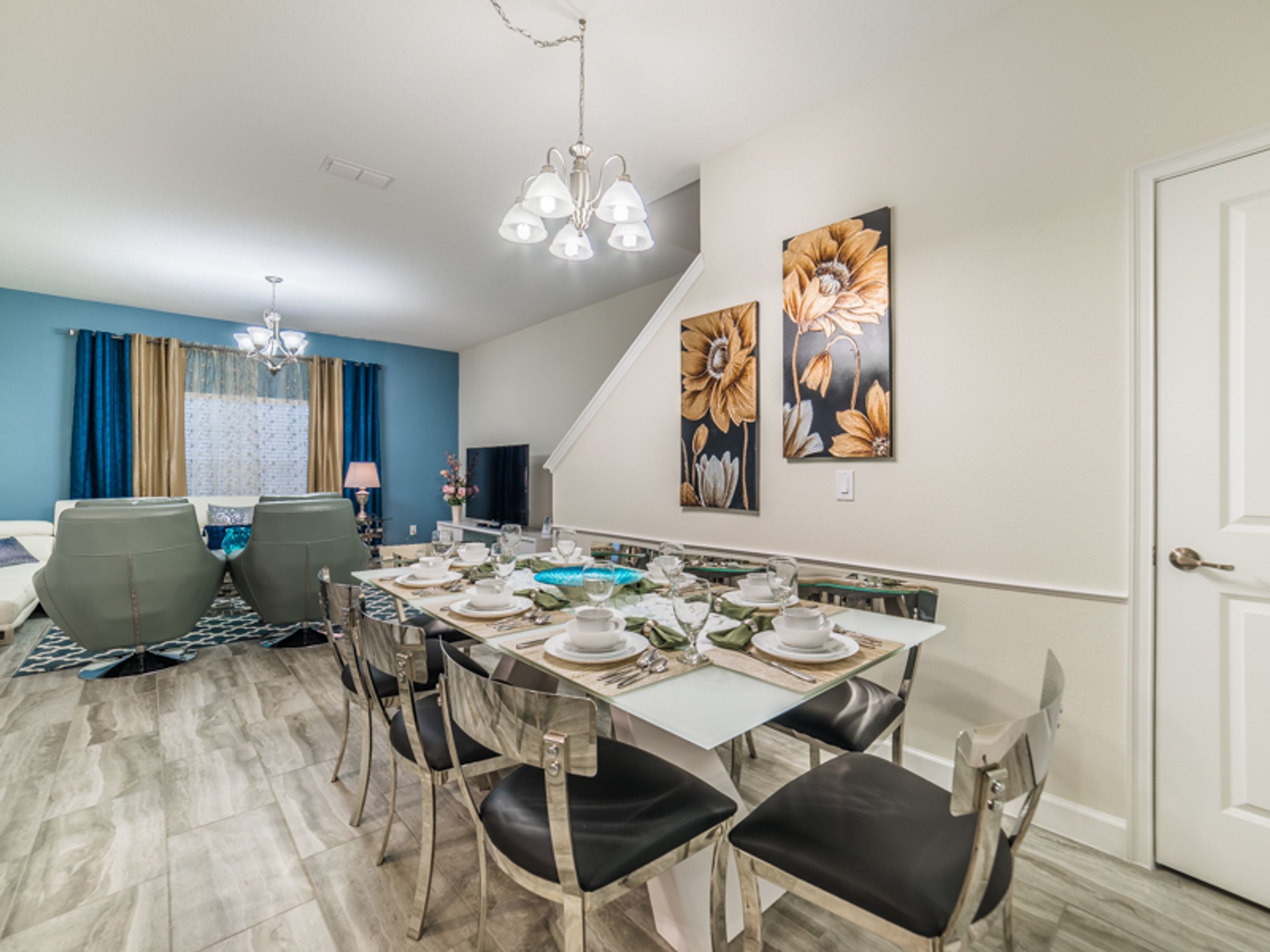 A dining area with a long table set for six, surrounded by chairs. There is a chandelier above the table, and artwork on the walls.