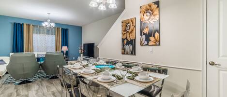 A dining area with a long table set for six, surrounded by chairs. There is a chandelier above the table, and artwork on the walls.