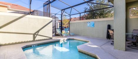 A swimming pool with a glass enclosure, lounge chairs, and a patio area with tables and chairs.