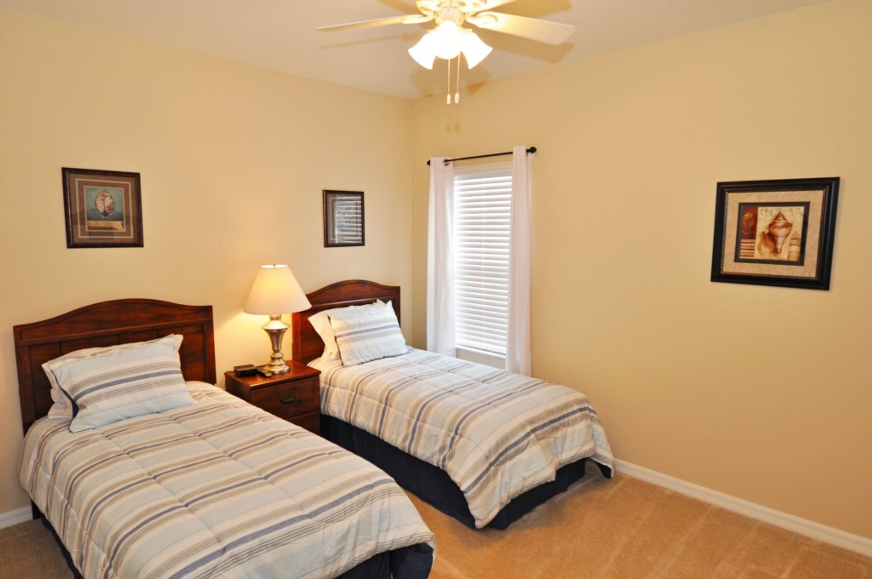 A bedroom with two beds, a ceiling fan, a window with blinds, and framed pictures on the walls.
