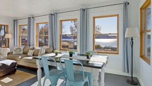 Ein geräumiges Wohnzimmer mit einem großen Fenster mit Blick auf einen malerischen See und Berge. Es gibt einen Essbereich mit Tisch und Stühlen sowie eine gemütliche Sitzecke mit einer Couch und Kissen.