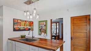 Eine Küche mit Holzausziehtisch, Spüle aus Edelstahl und einer Pendelleuchte. An der Wand hängt ein farbenfrohes Bild und ein Korkbrett mit verschiedenen Papieren.