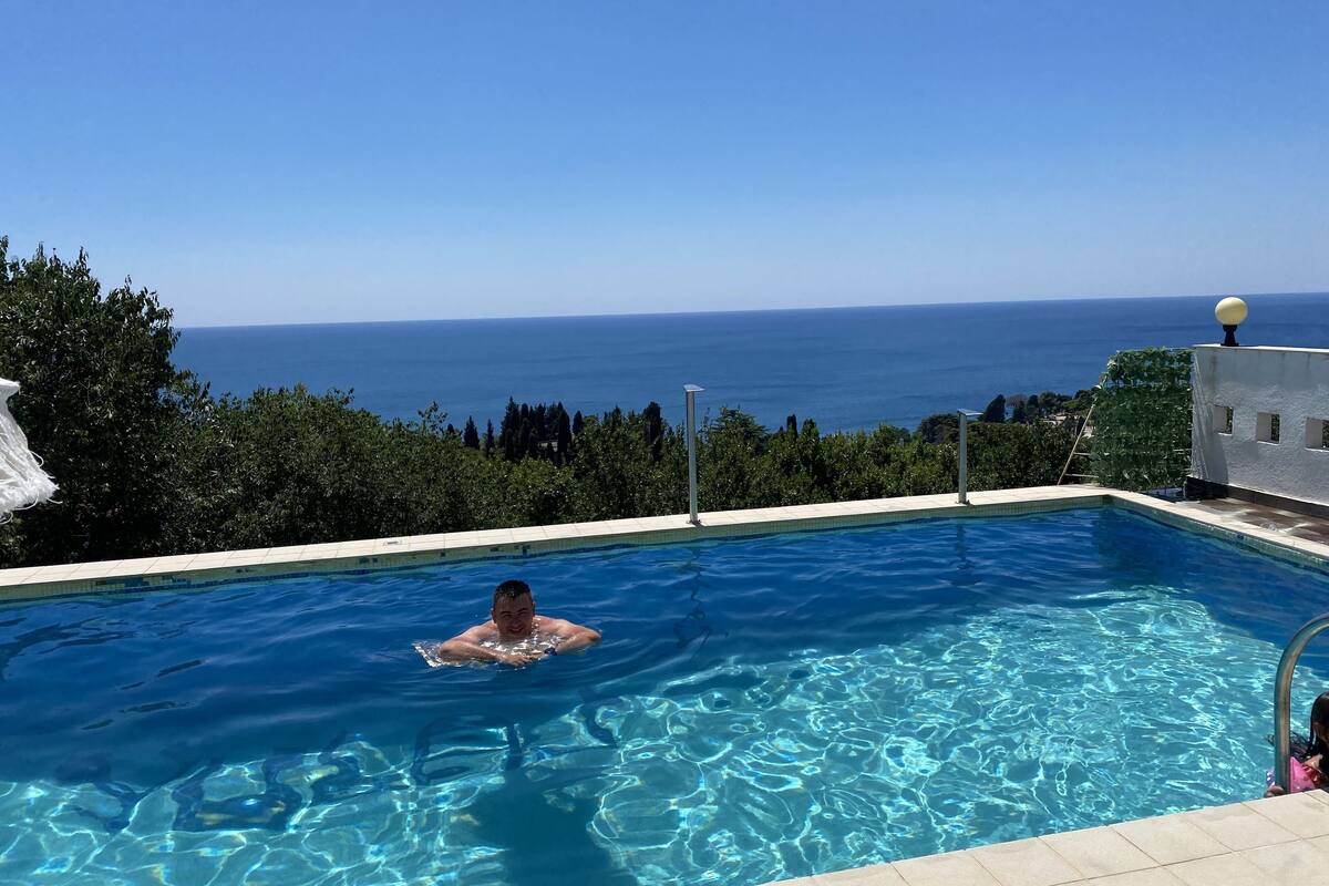 A swimming pool with clear blue water, surrounded by greenery and overlooking a scenic view of the sea.