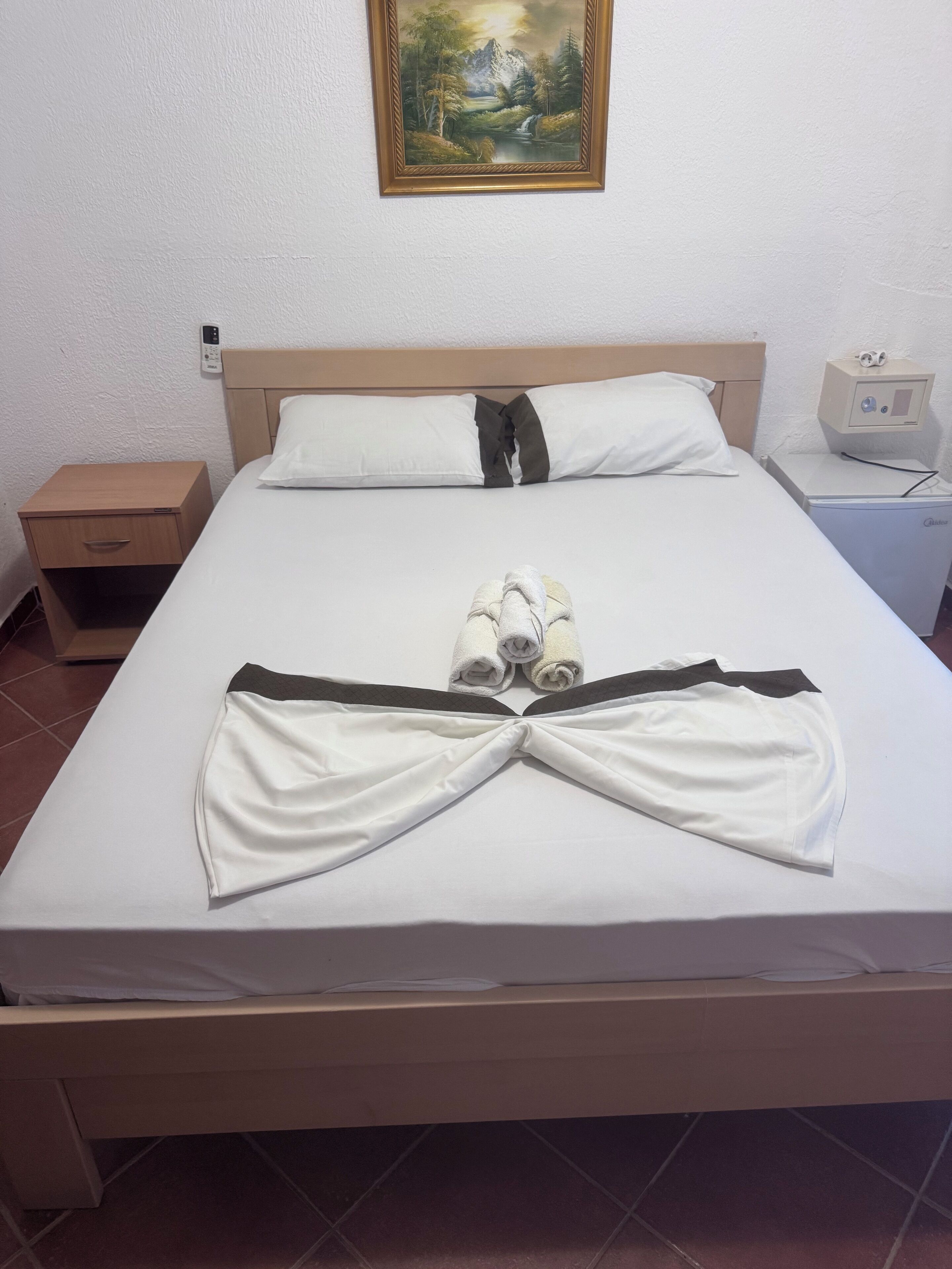 Una cama bien hecha con toallas dispuestas en forma de lazo, una mesita de noche con lámpara, un atril y un cuadro en la pared.