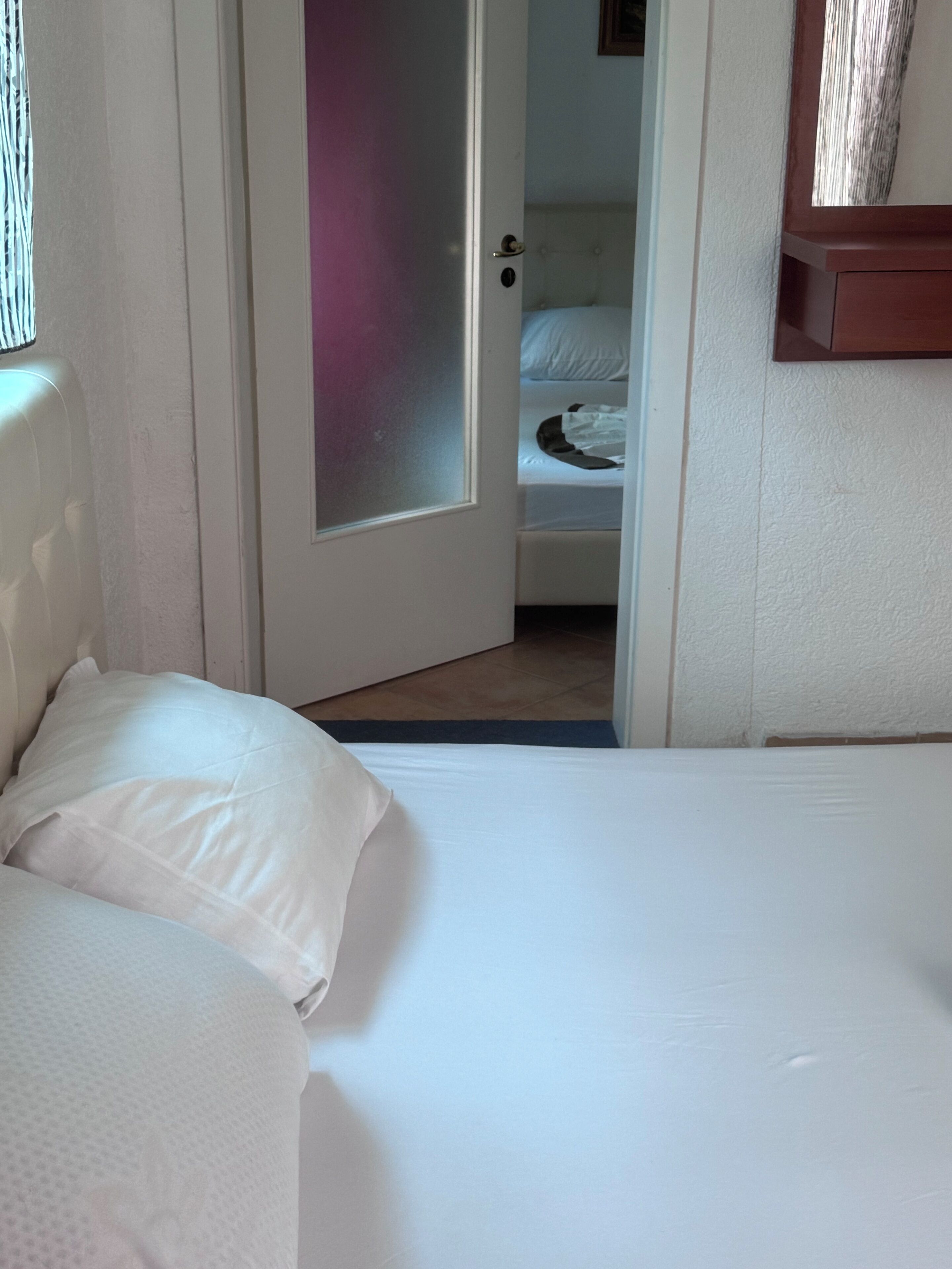 Une chambre d’hôtel avec un lit, une porte donnant sur une salle de bain et une petite étagère en bois au-dessus du lit.