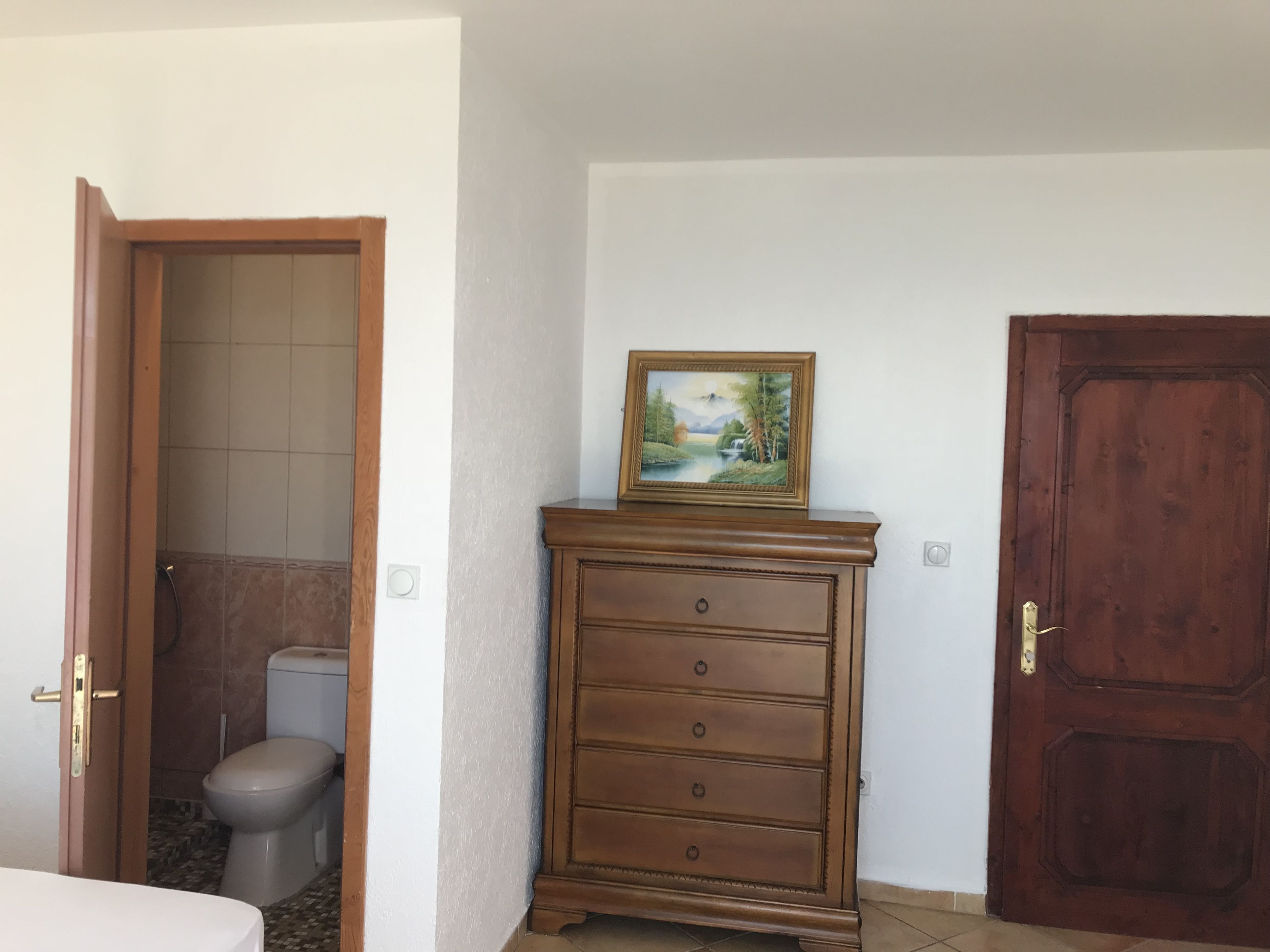 A bathroom with a toilet, a shower, and a wooden dresser with a painting.