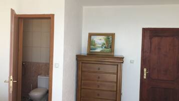 A bathroom with a toilet, a shower, and a wooden dresser with a painting.