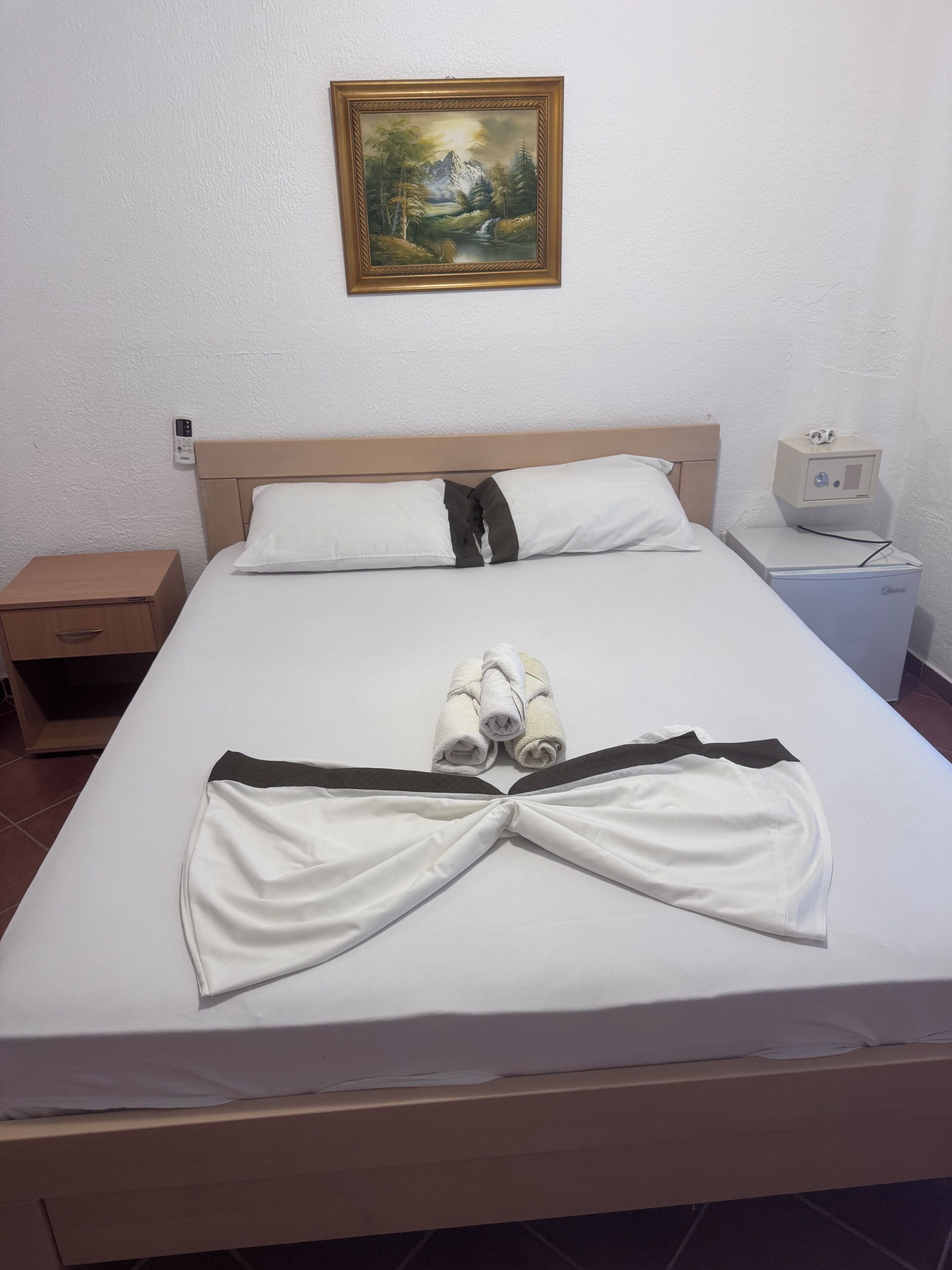 A neatly made bed with towels arranged in a bow, a bedside table, a nightstand with a lamp, and a painting on the wall.
