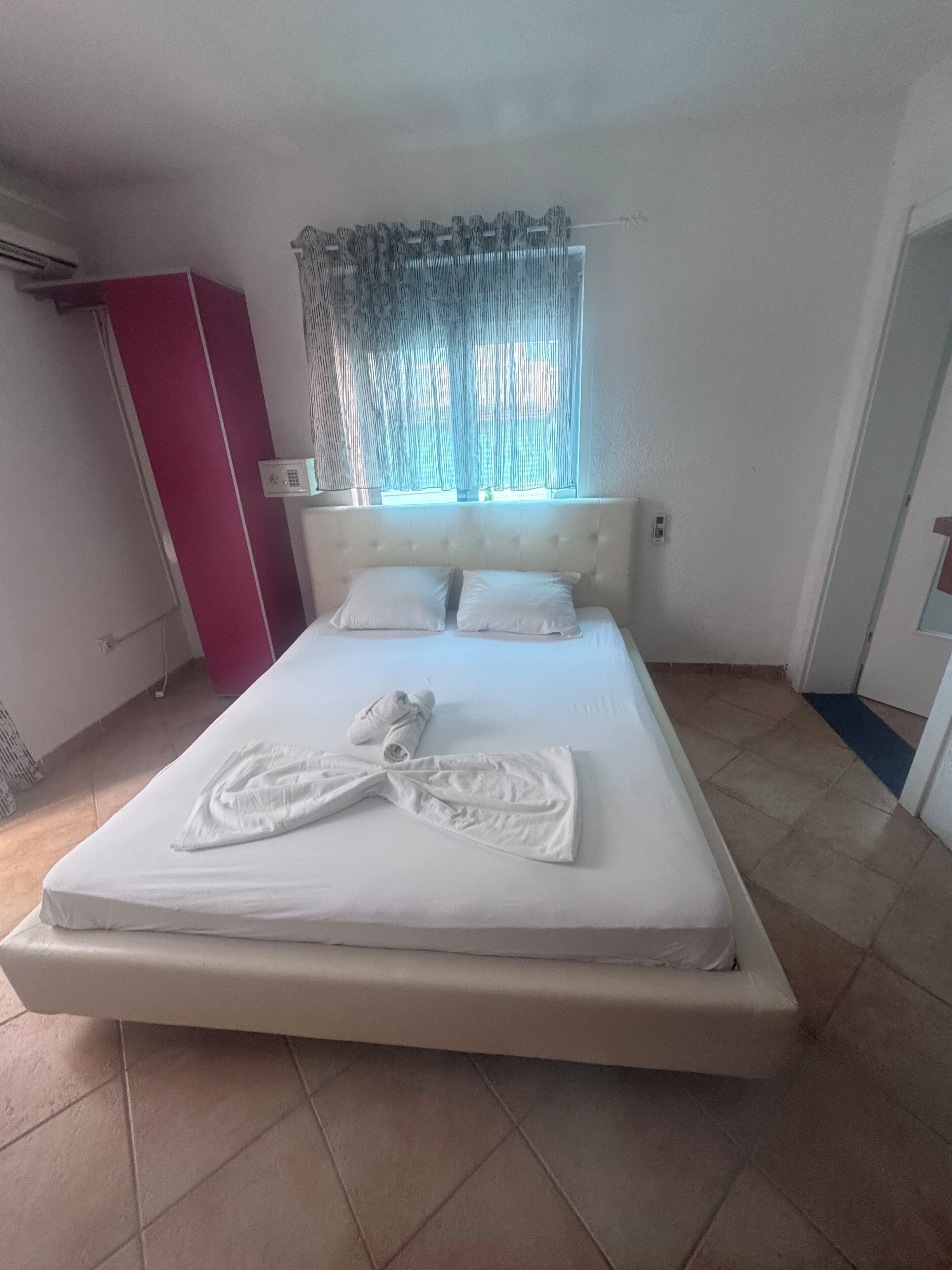 A neatly made bed with white linens and towels, a window with sheer curtains, a red wardrobe, and a white air conditioning unit.