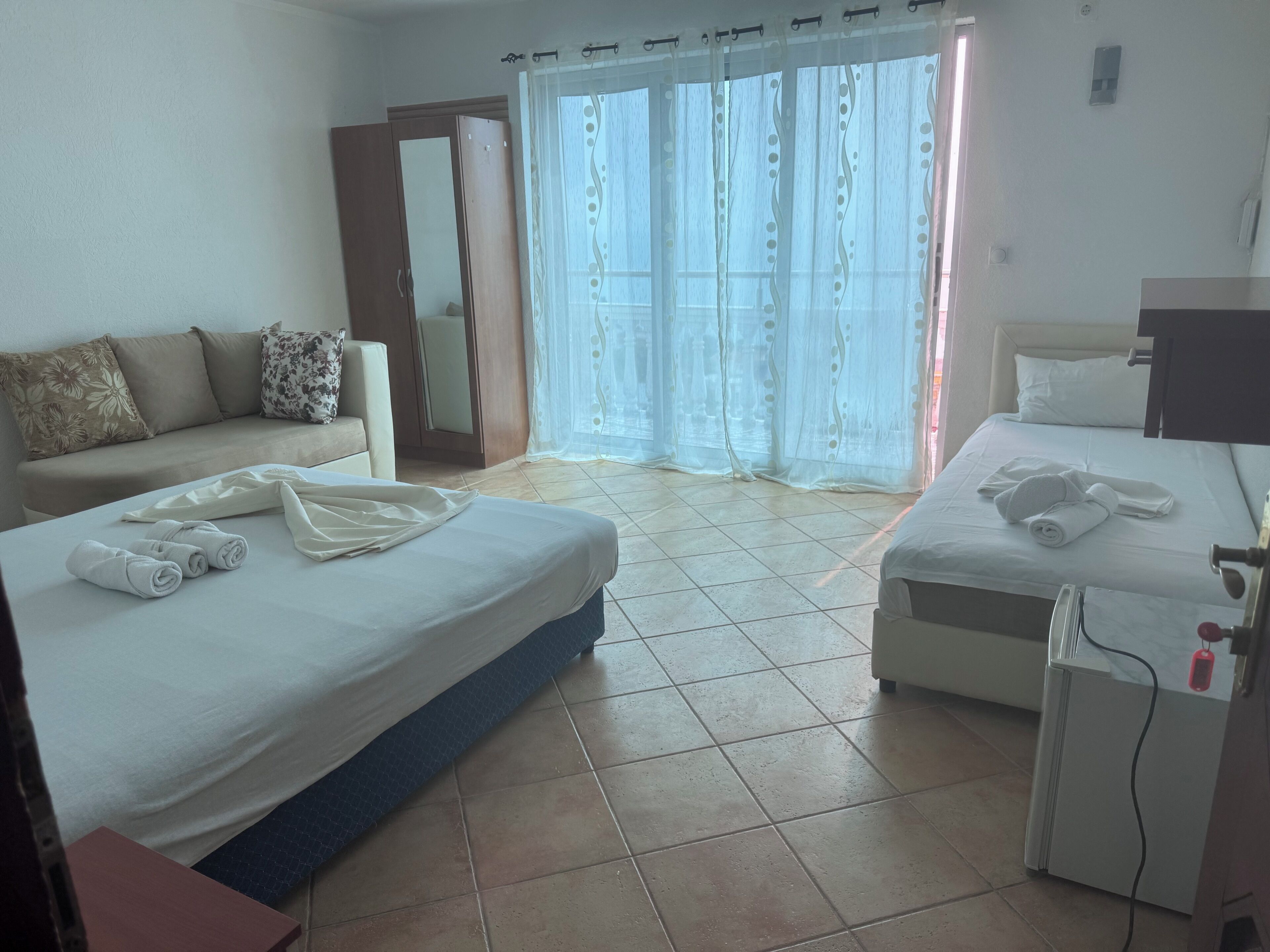 A hotel room with two beds, a sofa, a desk, and a balcony with curtains.