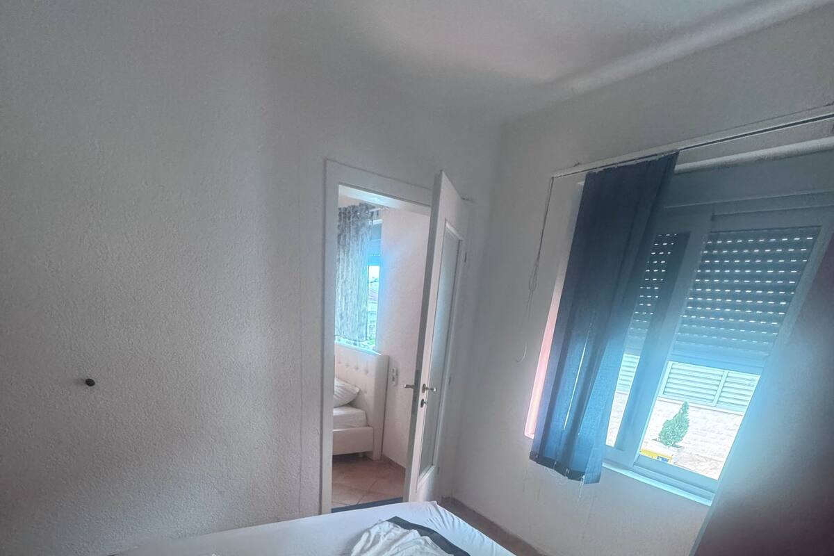 A neatly made bed with a white bedspread and two rolled towels, a window with blinds, and a door leading to a room with a white chair and a table.