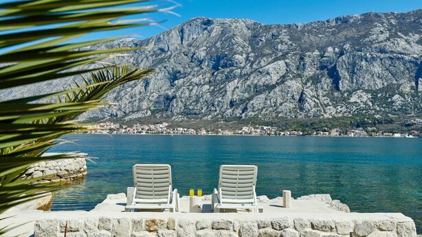 A seaside terrace with two white lounge chairs and a table, overlooking a calm sea and a mountainous backdrop.
