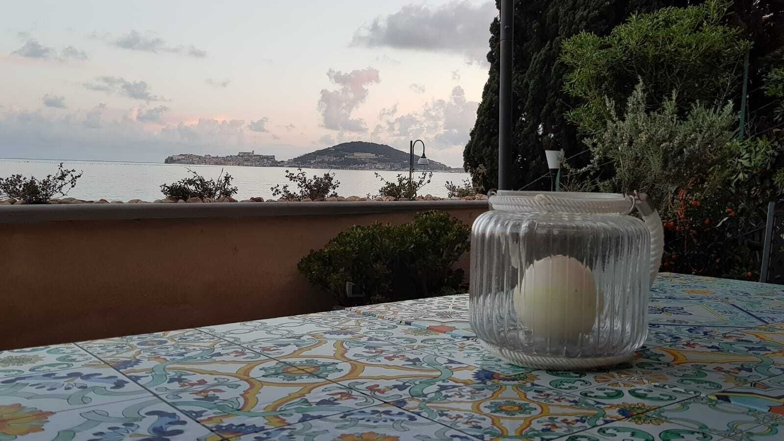 Une table recouverte d’une nappe à carreaux aux couleurs vives, un bocal en verre transparent contenant un objet blanc, et une vue sur une étendue d’eau avec une île lointaine et des bâtiments.