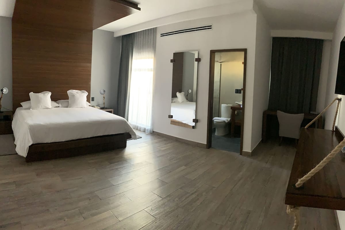 A hotel room with a large bed, a wooden floor, a mirror, a small bathroom, and a rope swing.