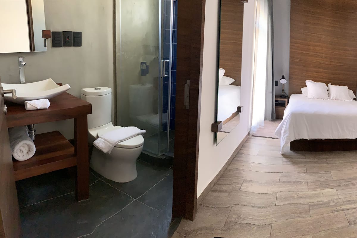 A bathroom with a sink, toilet, and shower. The room has a wooden floor and a bed with white bedding.