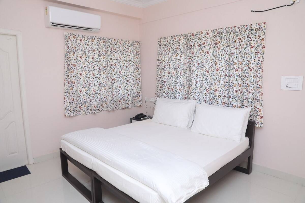 A bedroom with a bed, floral curtains, an air conditioning unit, and a bedside table.