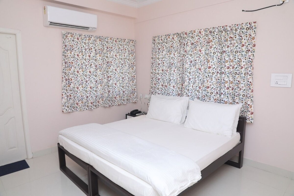 A bedroom with a bed, floral curtains, an air conditioning unit, and a bedside table.