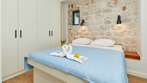 Une chambre avec un mur en pierre, un lit recouvert d’une couette bleue, des oreillers blancs et une serviette ornée de motifs floraux. On y trouve deux appliques murales noires et une petite fenêtre.
