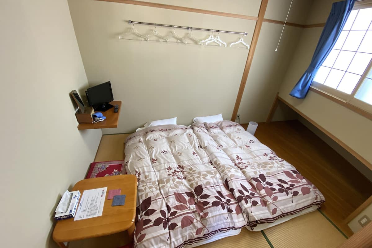 Een keurig opgemaakt hotelkamer met een bed, een nachtkastje met een lamp, een klein plankje met persoonlijke spullen en een raam met gordijnen.