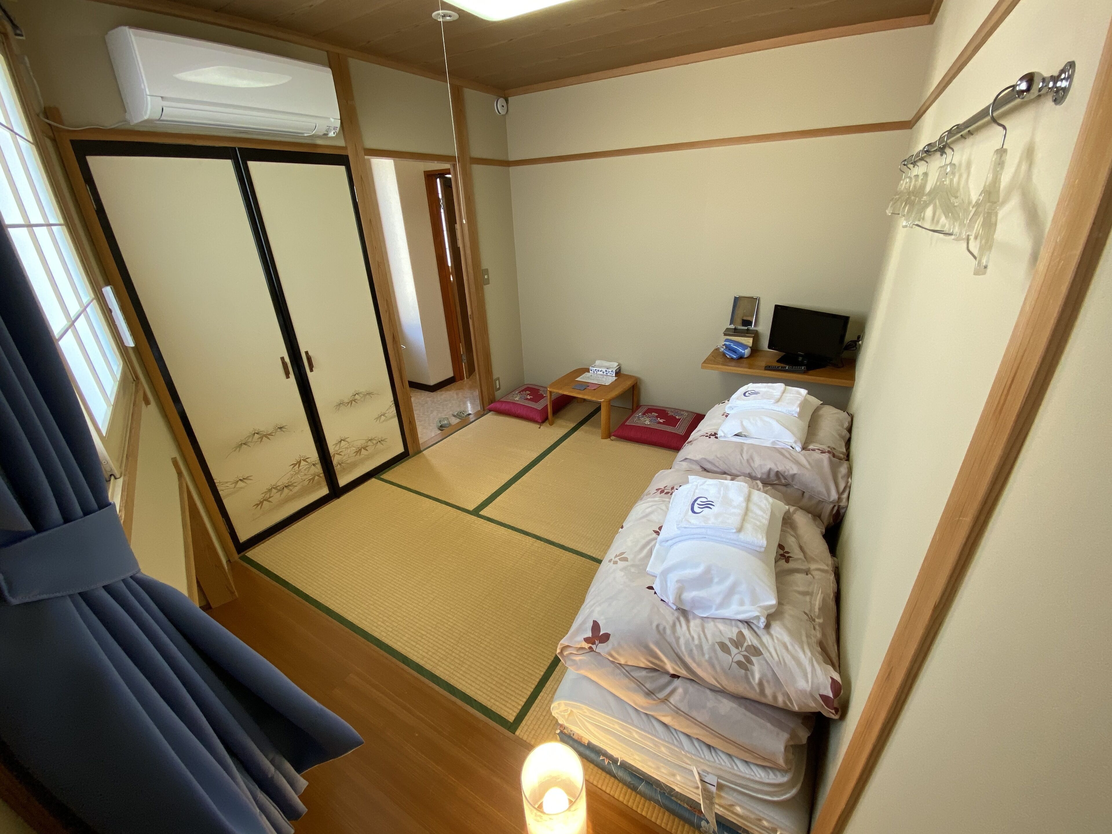 Una habitación japonesa tradicional con suelo de tatami, futón, una pequeña mesa con lámpara y televisión.
