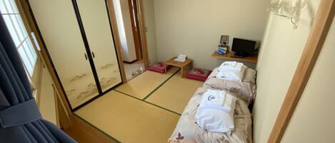 Una habitación japonesa tradicional con suelo de tatami, futón, una pequeña mesa con lámpara y televisión.