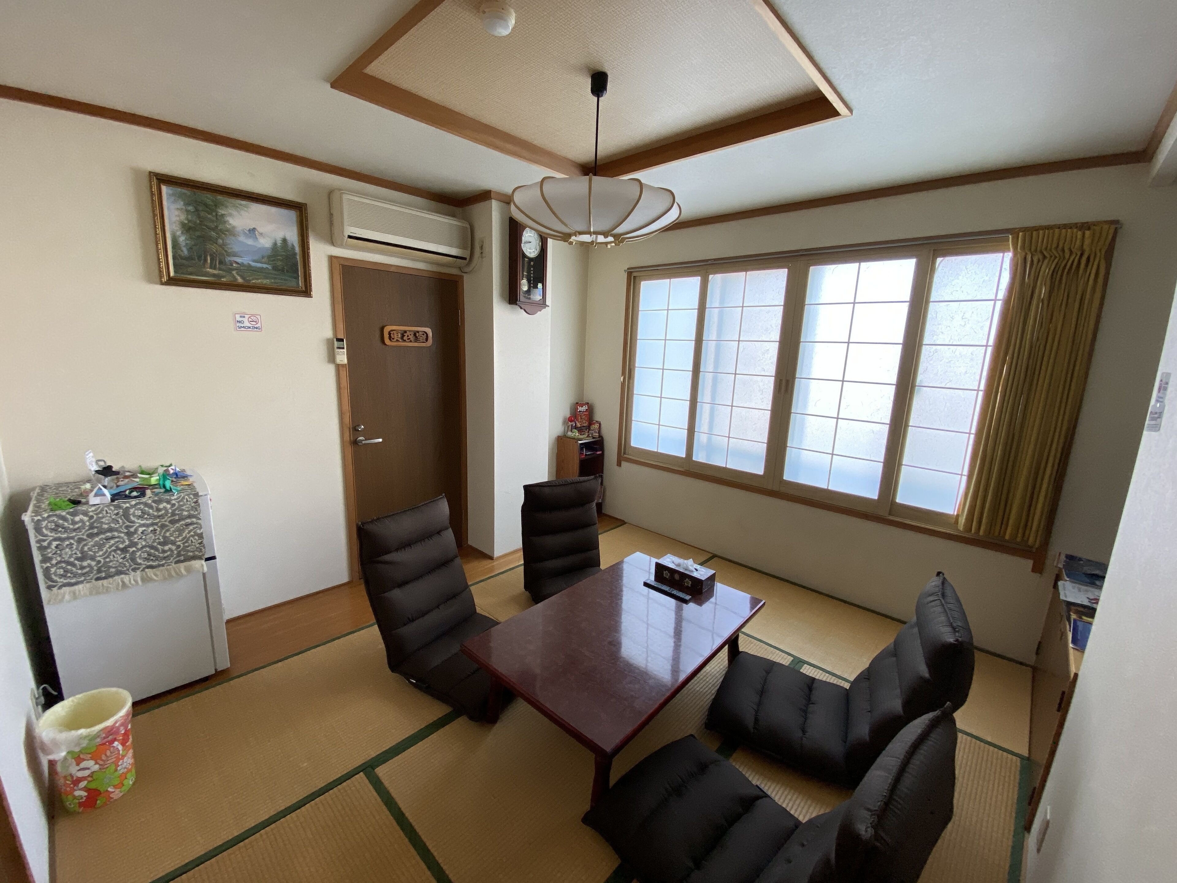 Quarto com piso de tatame, uma mesa de madeira com cadeiras, geladeira, ar-condicionado, relógio e uma janela grande com cortinas.