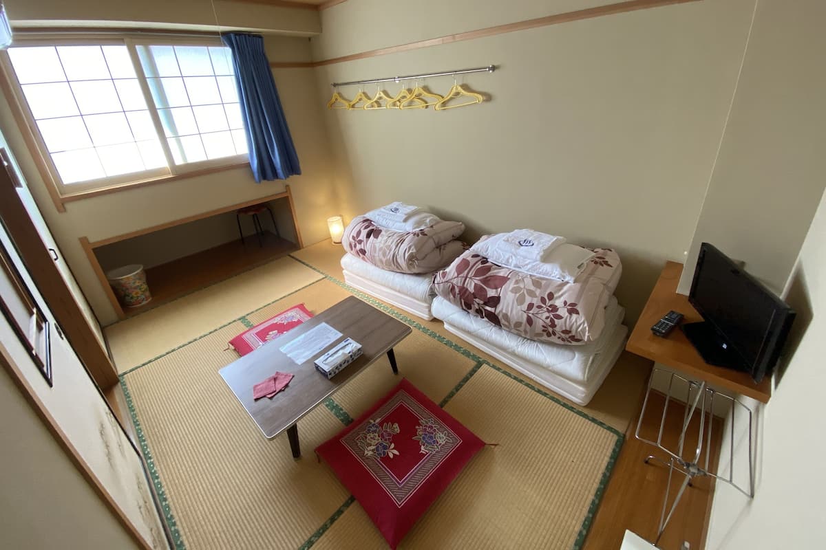 Een compacte, verzorgde kamer met een bed, een televisie, een tafel met een rode mat en een raam met gordijn.