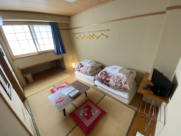 A small, neatly arranged room with a bed, a TV, a table with a red mat, and a window with a curtain. - Nozawa Dream Central (Nozawaonsen)