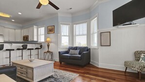 Uma sala de estar espaçosa com piso de madeira, um sofá cinza, uma poltrona estampada, um armário branco e um ventilador de teto.