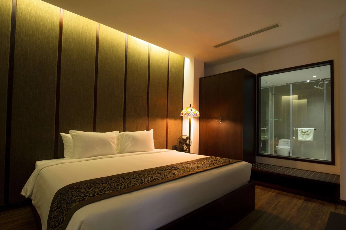 A hotel room with a large bed, a nightstand with a lamp, a wardrobe, and a glass shower enclosure.