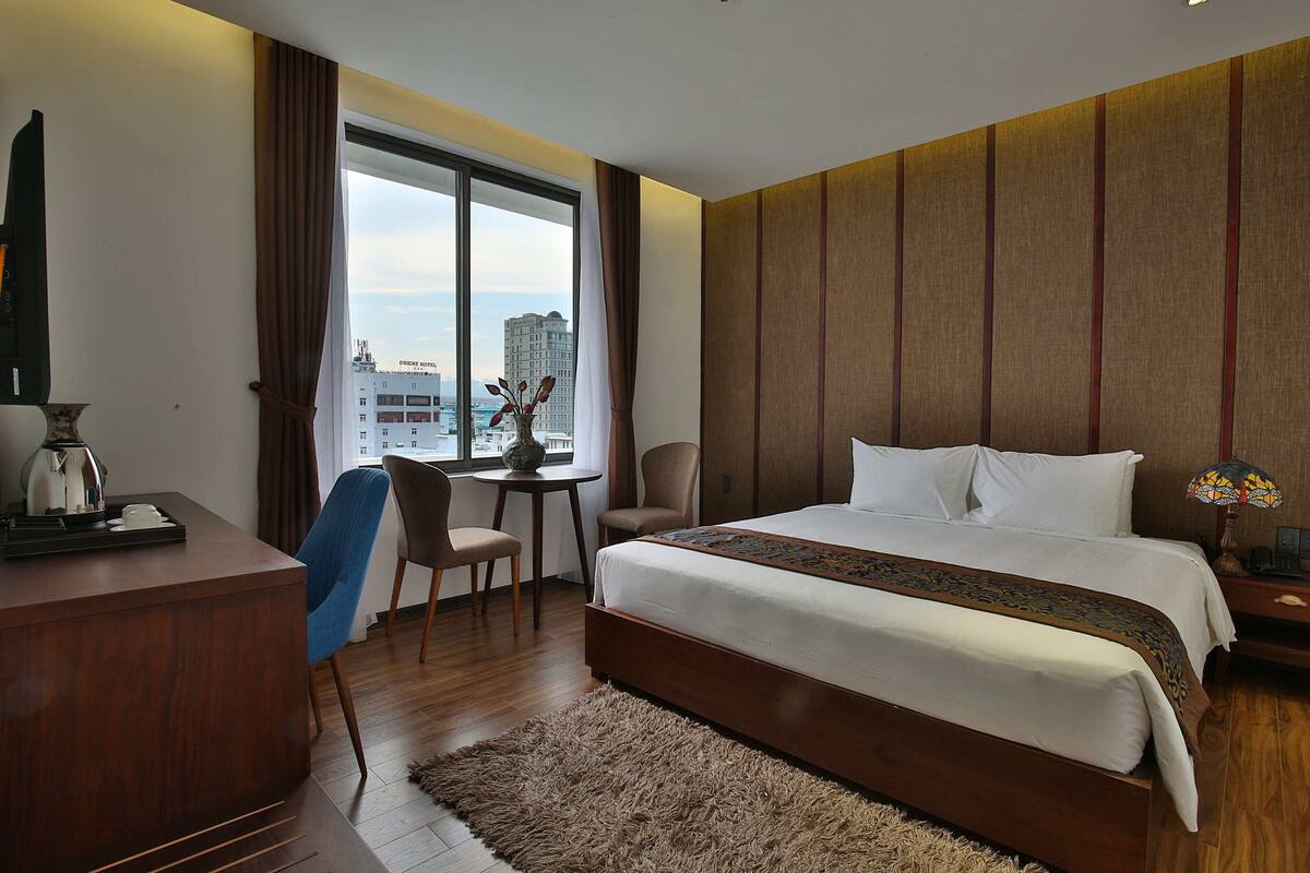 A hotel room with a large bed, a desk with a coffee maker, a chair, a small table with a vase, and a window with a view of buildings.