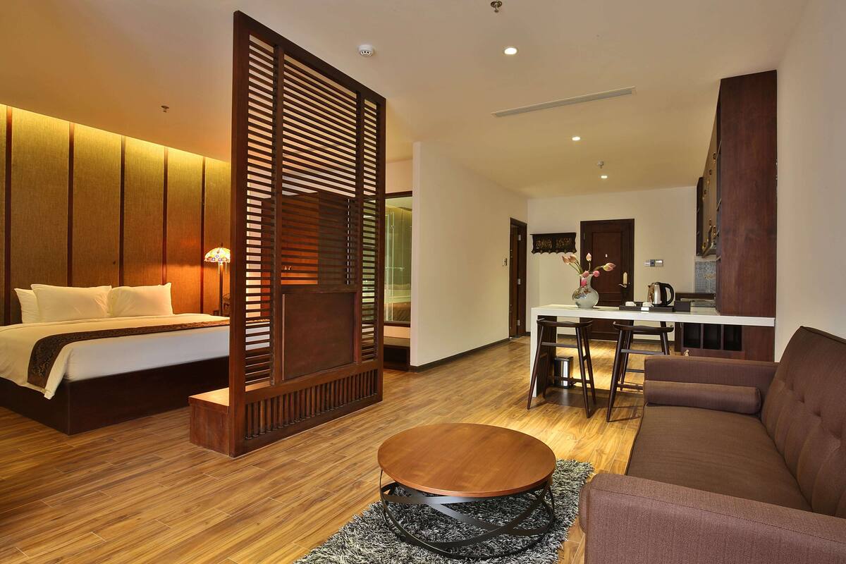 A modern hotel room with a bed, a sofa, a round coffee table, and a dining area with a table and chairs.