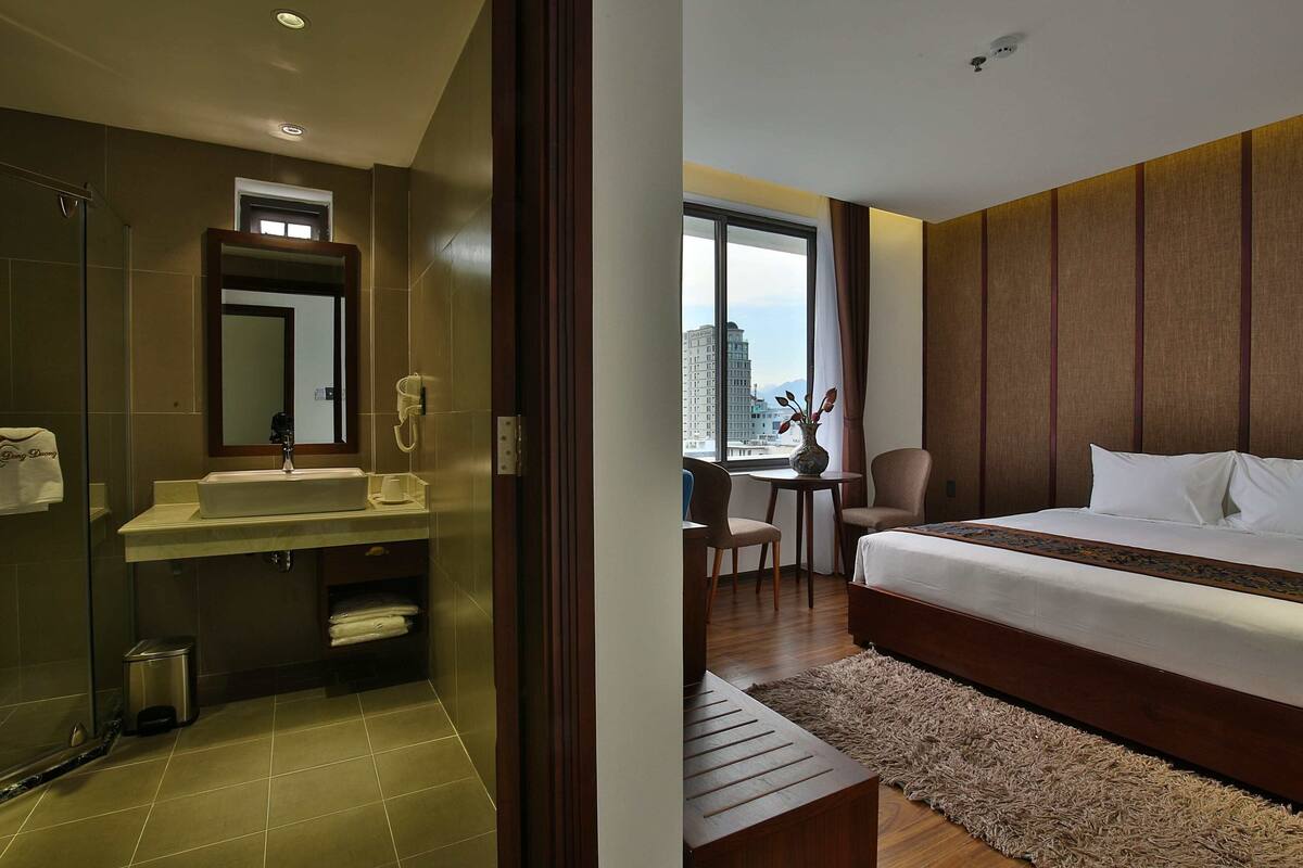 A hotel room with a bathroom, a bed, a desk, and a view of the city.