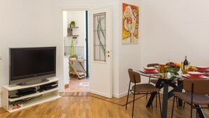 A modern living room with a flat-screen TV, wooden flooring, a dining area with a glass table and chairs, and a door leading to a kitchen. - Bright, stylish, renovated Flat near Villa Borghese (Roma)