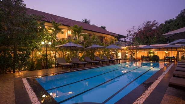 A swimming pool with lounge chairs and umbrellas, surrounded by a landscaped garden and a building with a red-tiled roof. - RATA  Boutique (Siem Reap)
