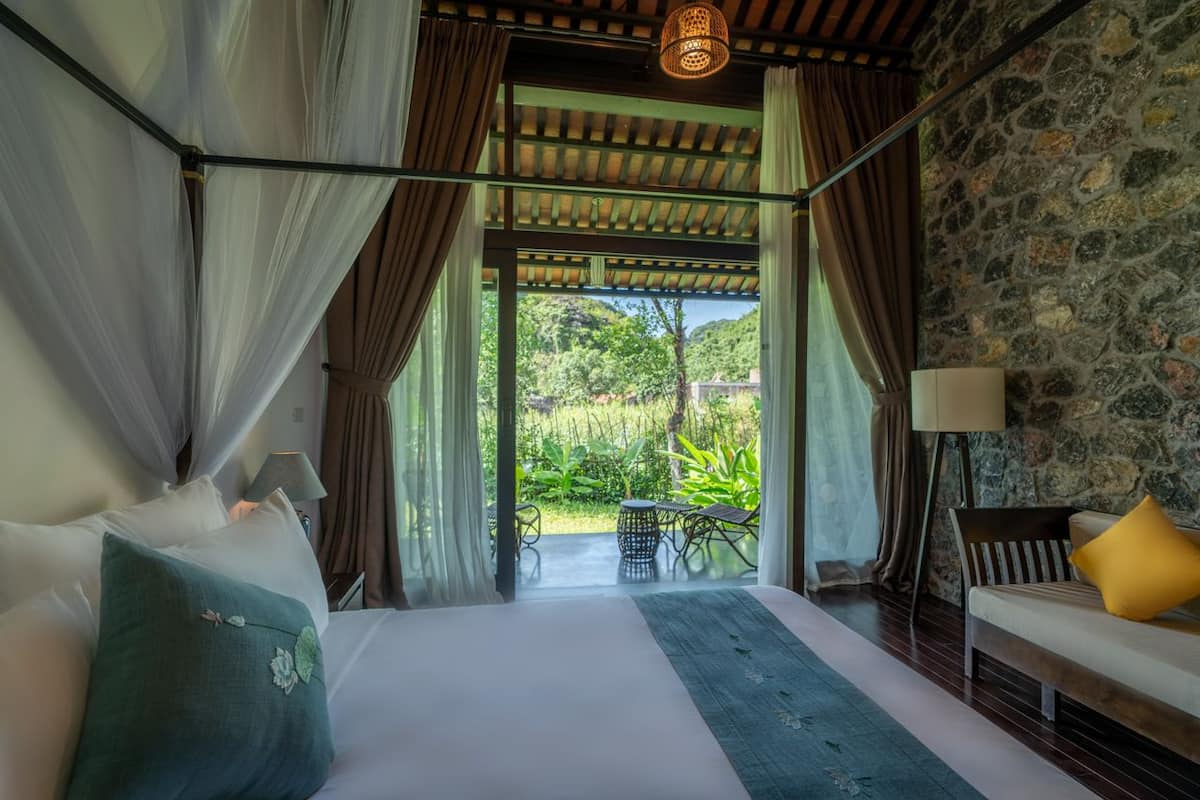 A bedroom with a bed, a bench, a lamp, and a view of the outdoors.