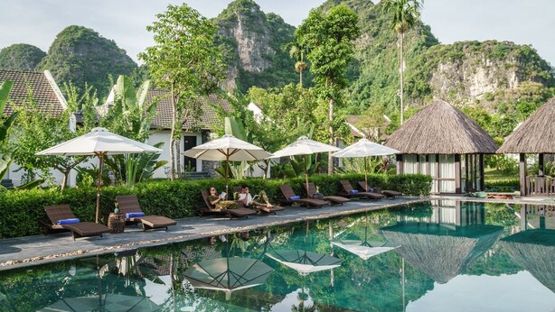 A poolside area with lounge chairs and umbrellas, surrounded by lush greenery and a mountainous backdrop. - Aravinda Resort (Nam Hoa Lu)